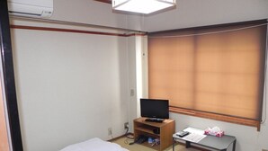 Ein kleines Hotelzimmer mit einem Einzelbett, einem Fernseher auf einem Holzständer, einem kleinen Tisch mit einer Fernbedienung und einem Fenster mit Jalousien.