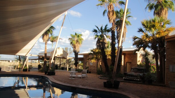 A pool area with palm trees, a brick patio, and a vintage car. - Longreach Outback Adventures (Longreach)