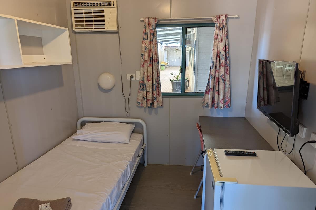 Una habitación pequeña con cama individual, escritorio, televisor, ventana con cortinas y aire acondicionado.