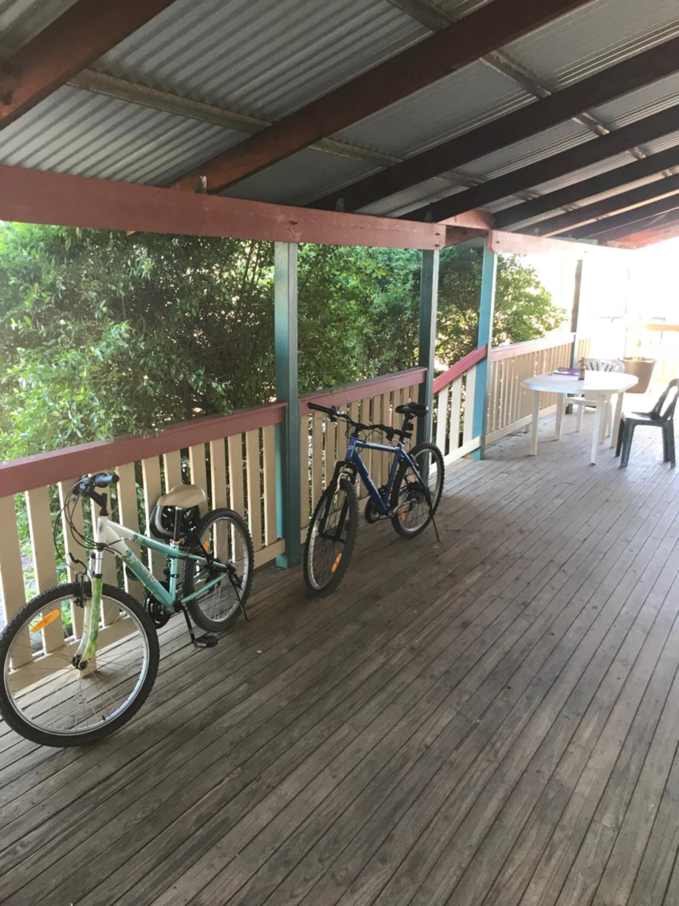 Una terraza cubierta de madera con dos bicicletas estacionadas, una mesa y sillas, y vistas a árboles al fondo.