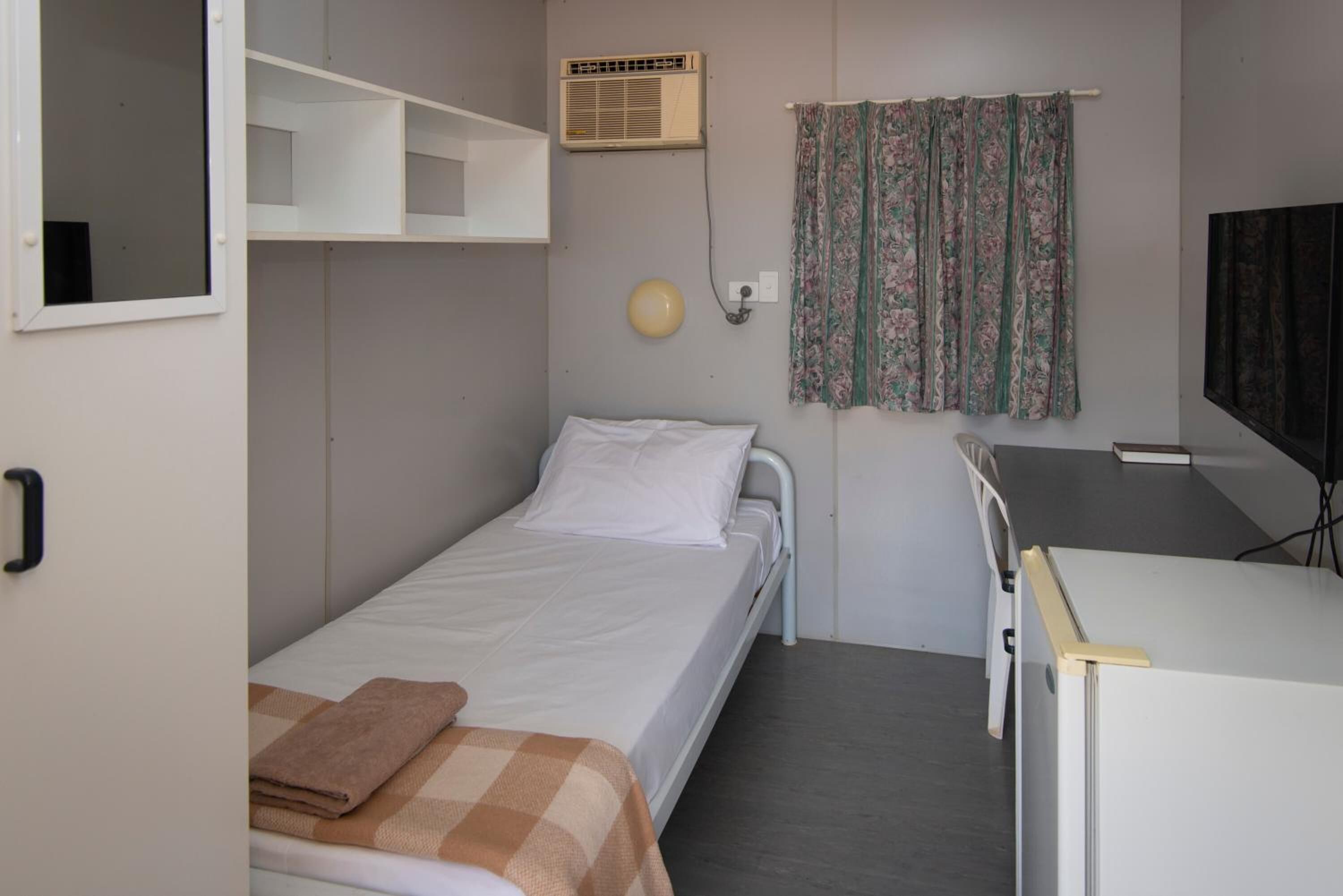 Ein kleiner, kompakter Raum mit einem Einzelbett, einem Schreibtisch, einem Stuhl, einem Kleiderschrank und einem Fernseher.