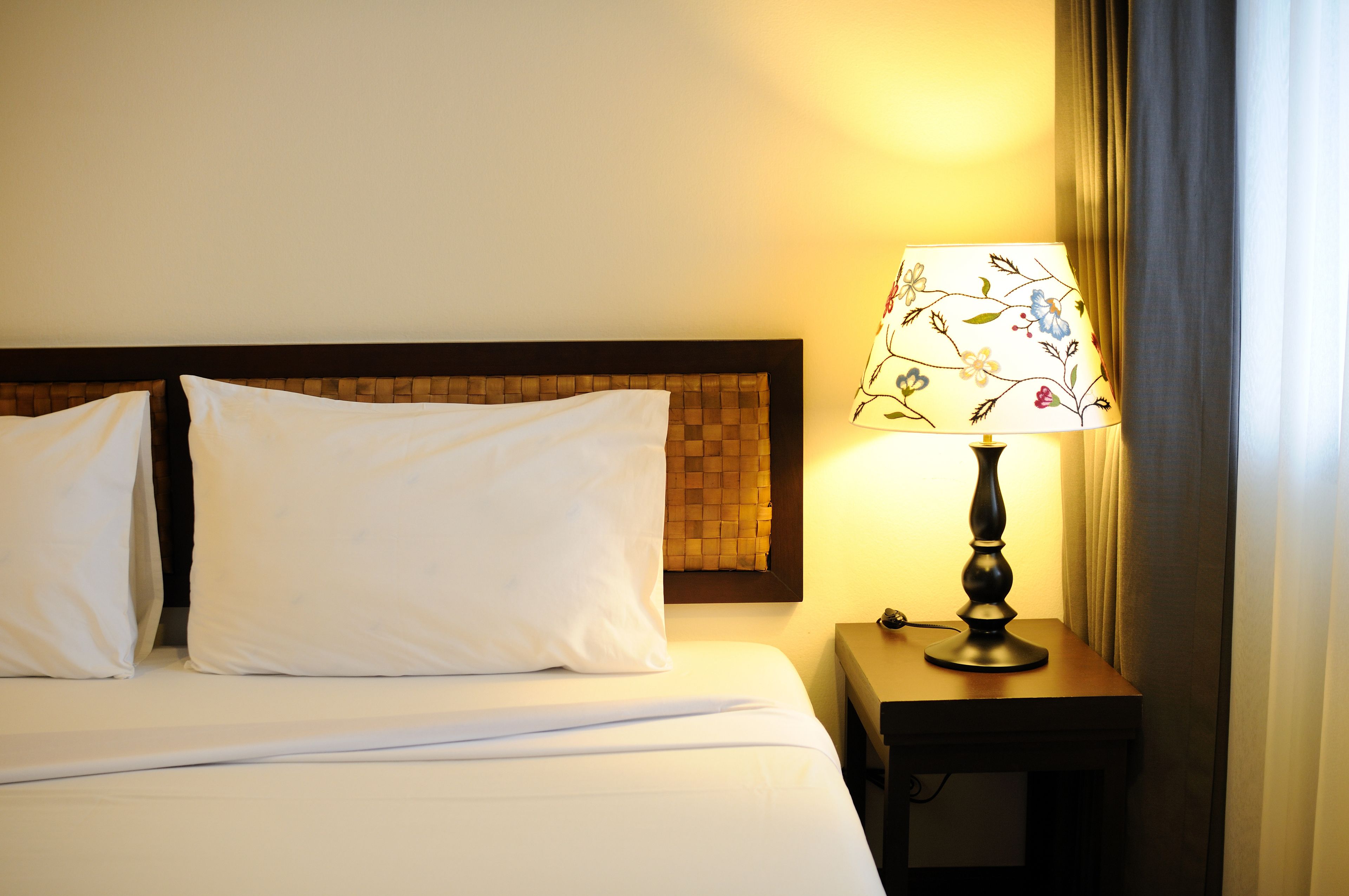 A neatly made bed with white linens and a wooden headboard. A table lamp with a floral design is placed on a wooden nightstand beside the bed.