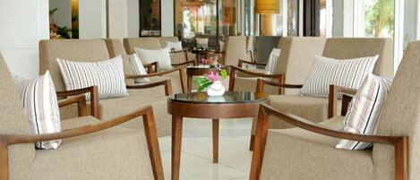 Un hall con sofás y sillones color beige, una mesa redonda de madera con un jarrón y flores, y ventanas amplias con vistas al exterior.