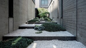 A narrow, stone-walled courtyard with a gravel path, large rocks, and green shrubs.