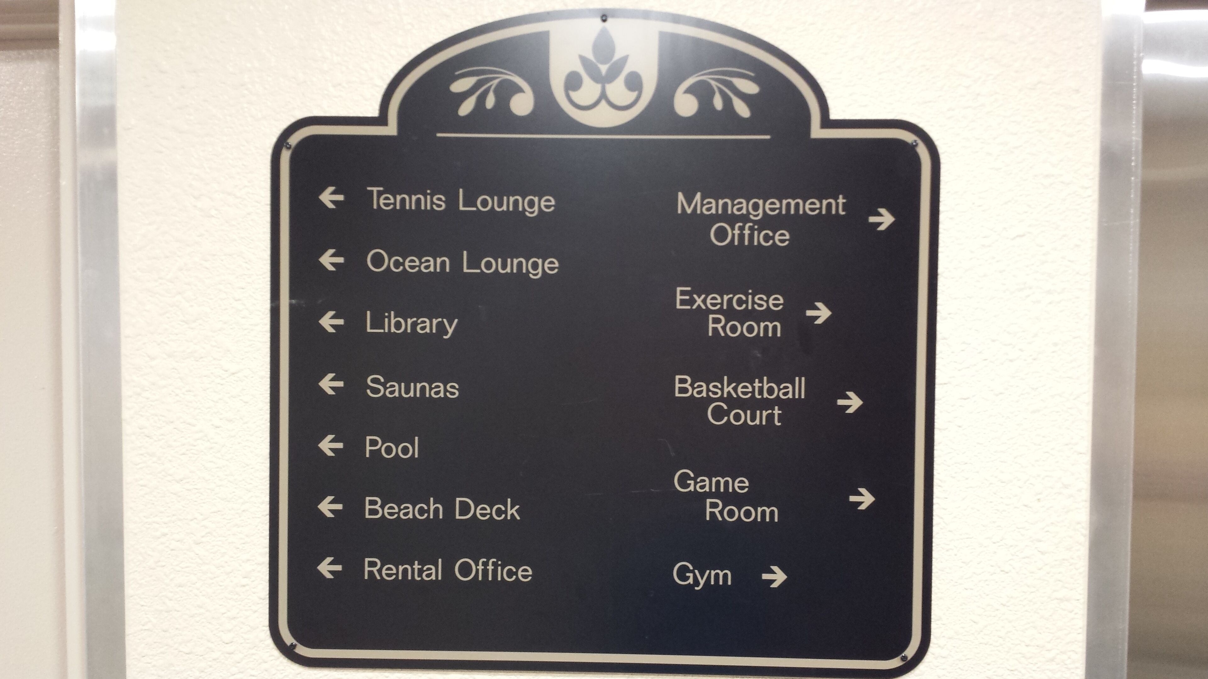 Een richtingwijzer met pijlen die wijzen naar diverse faciliteiten, zoals Tennis Lounge, Ocean Lounge, Bibliotheek, Saunas, Zwembad, Stranddeck, Verhuurbalie, Administratiekantoor, Fitnessruimte, Basketbalveld, Spelruimte en Gym.