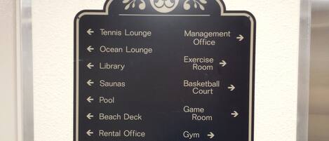 Een richtingwijzer met pijlen die wijzen naar diverse faciliteiten, zoals Tennis Lounge, Ocean Lounge, Bibliotheek, Saunas, Zwembad, Stranddeck, Verhuurbalie, Administratiekantoor, Fitnessruimte, Basketbalveld, Spelruimte en Gym.
