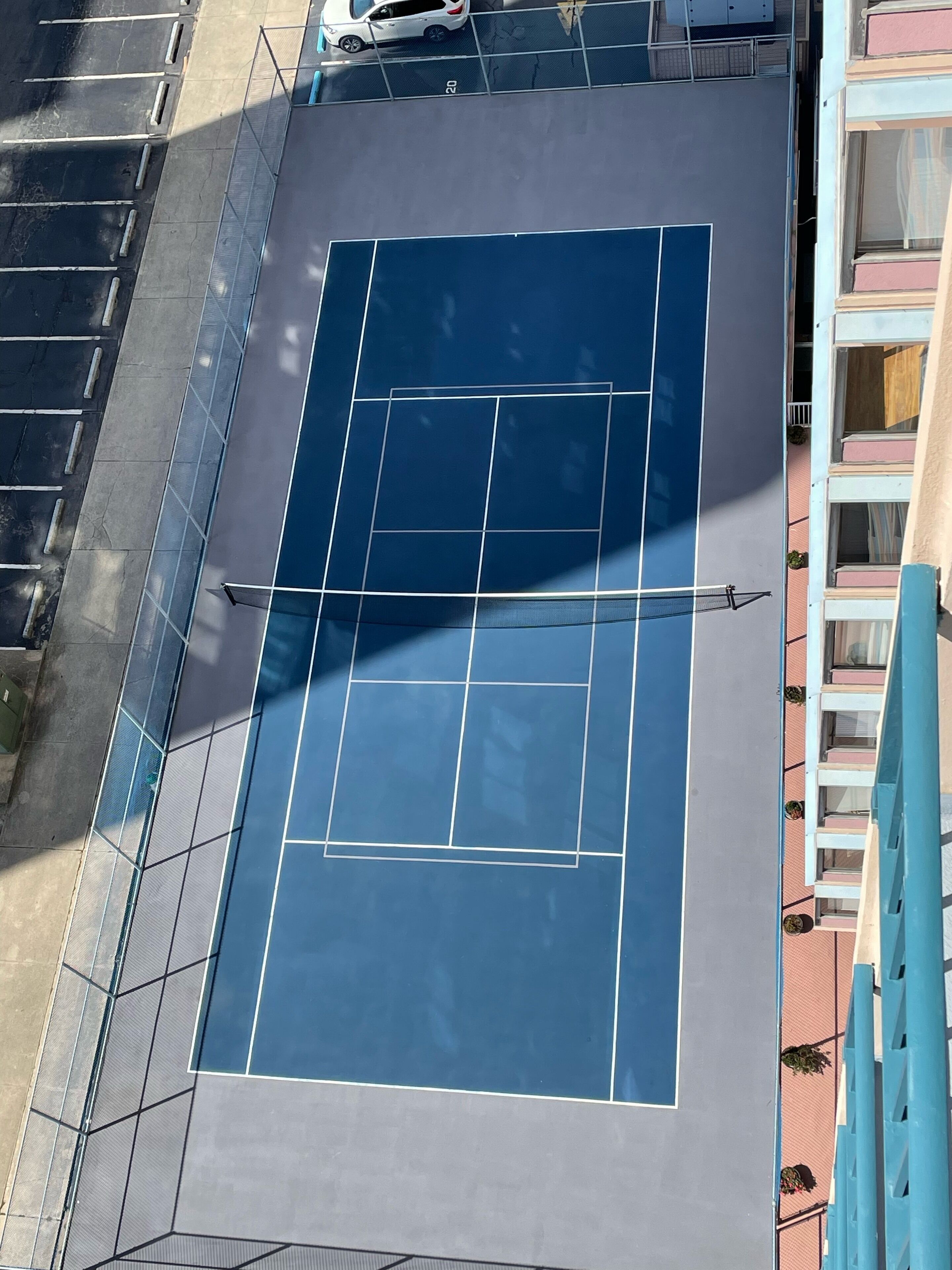Una cancha de tenis con una red, rodeada por una cerca de metal y un edificio con balcones.