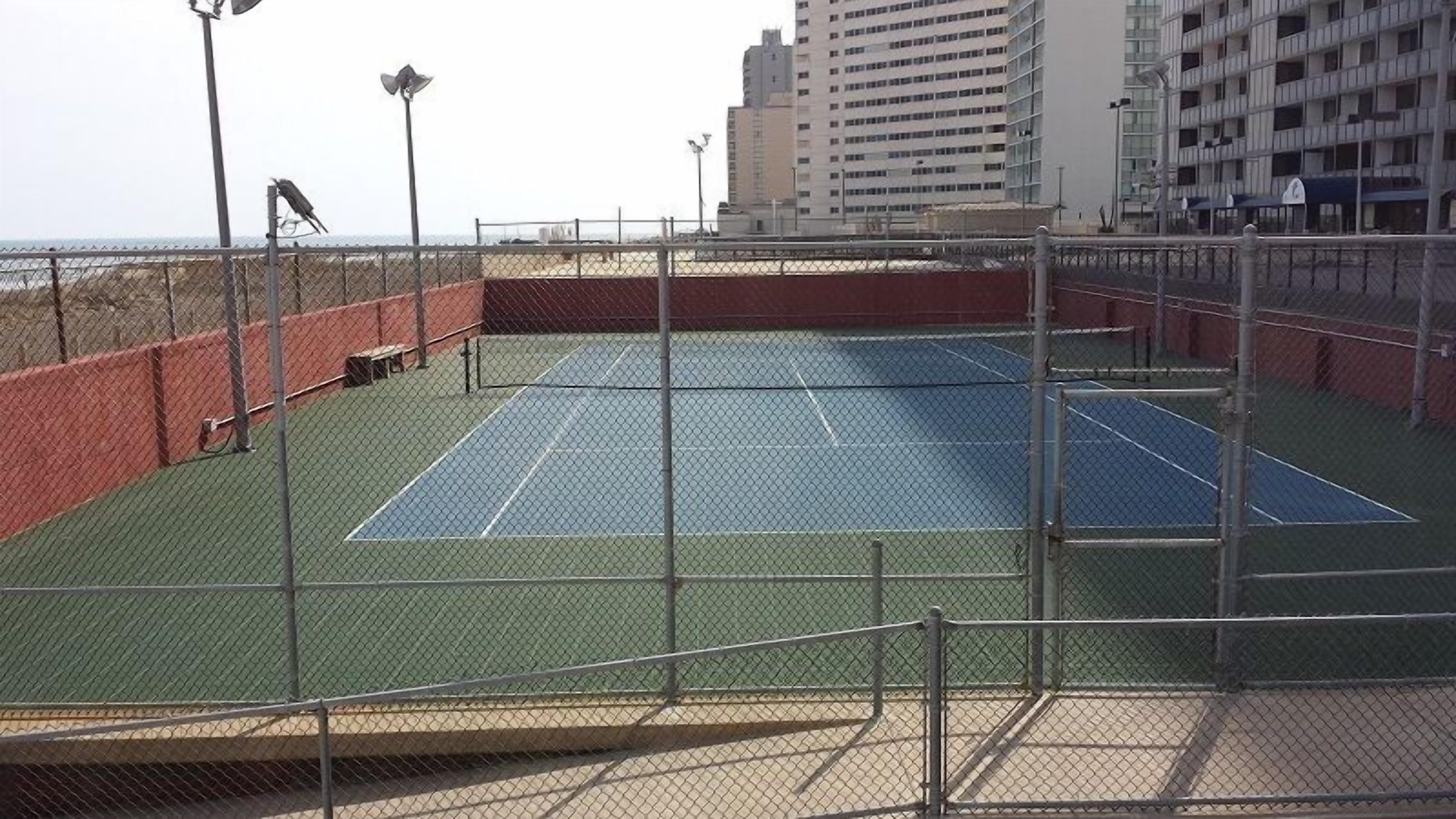 Una cancha de tenis al aire libre con una cerca de malla metálica, rodeada por un muro de ladrillo rojo y una baranda de metal.