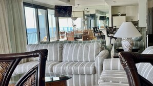 Ett modernt vardagsrum med en randig soffa, trästolar, ett soffbord i glas och utsikt över havet genom stora fönster.