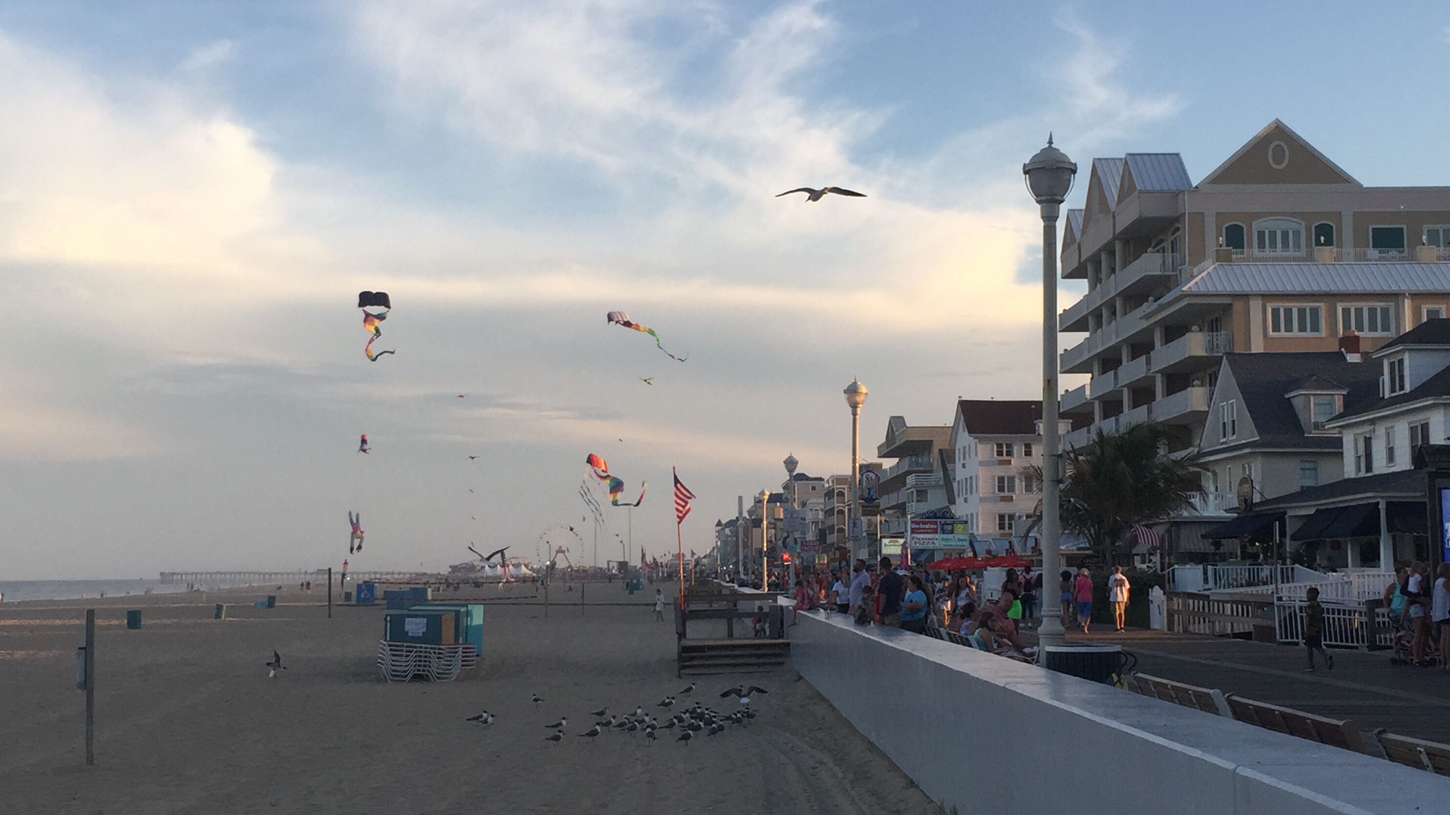 Une promenade en bord de mer bordée de bâtiments, une jetée visible au loin et des cerfs-volants qui volent dans le ciel.