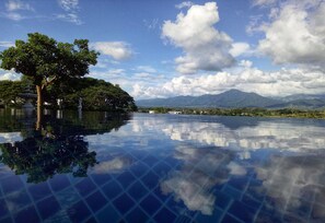 Uma área de piscina ao ar livre tranquila, com reflexo límpido do céu e das montanhas, rodeada por vegetação exuberante e uma fonte.