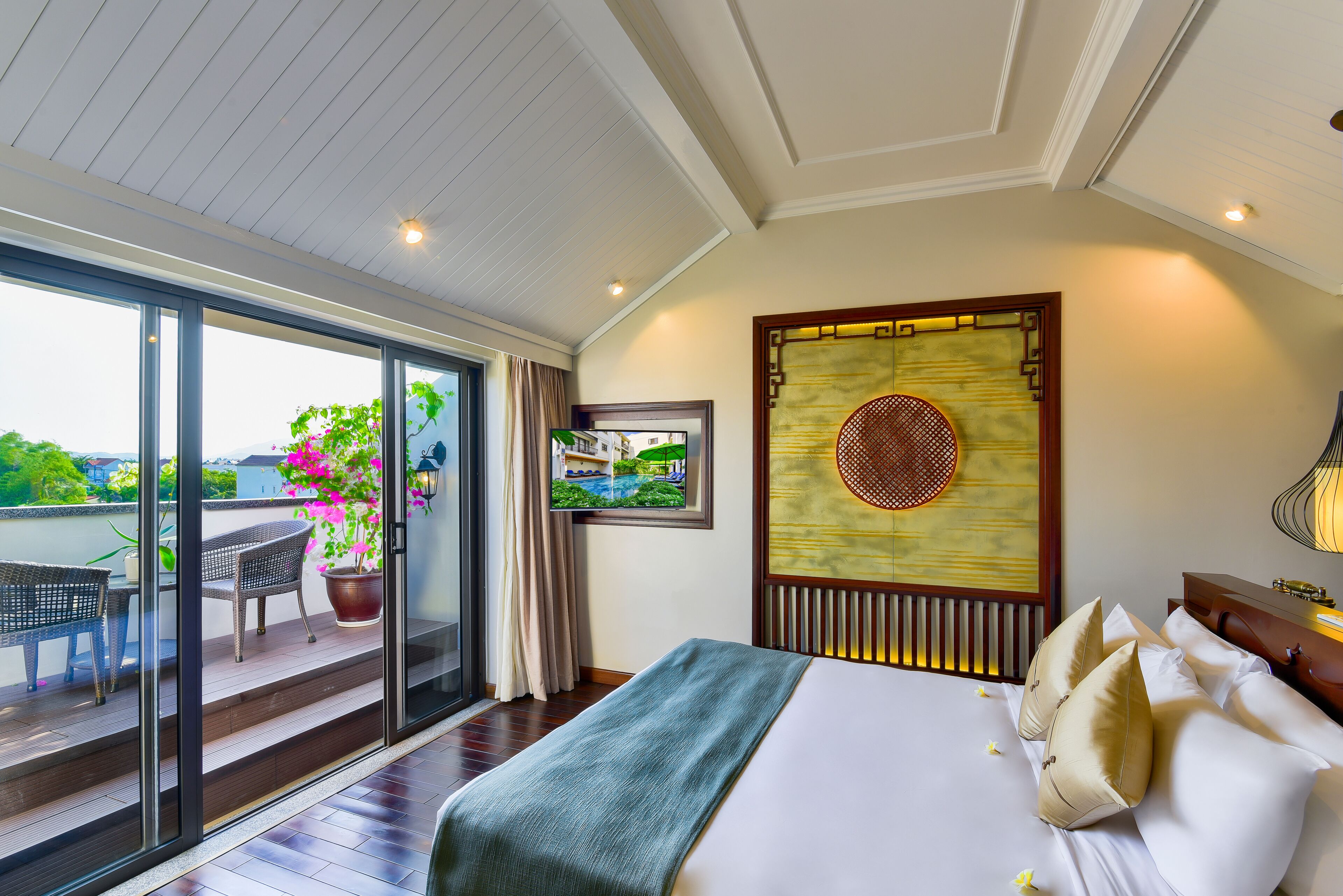 A hotel room with a balcony, a bed with pillows, a wall-mounted artwork, and a television.
