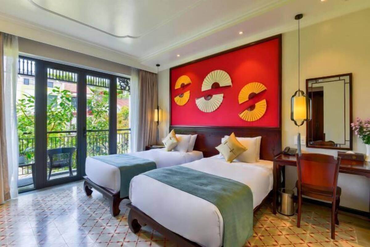 Habitación de hotel con dos camas, un ventanal con vistas a zonas verdes, una pared de acento roja y un escritorio con una silla.