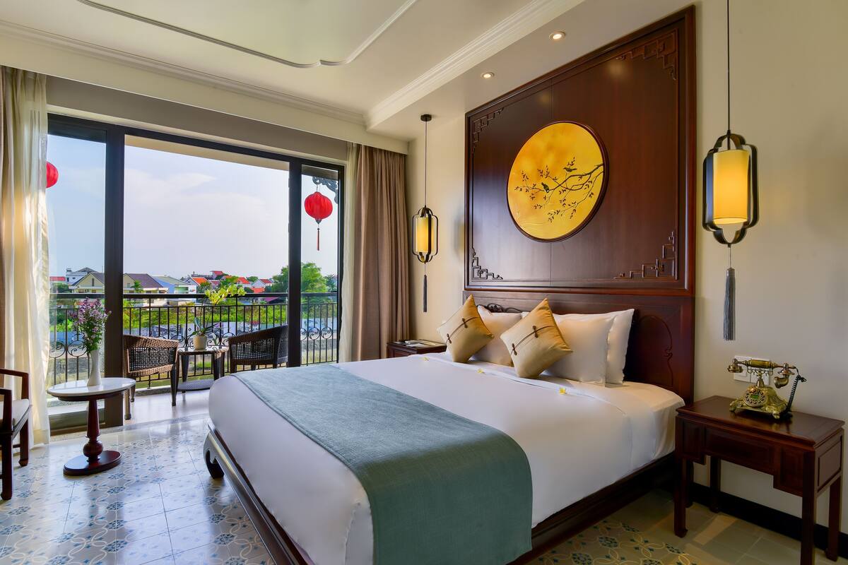 Habitación de hotel con una cama grande, balcón con mobiliario exterior y vistas del entorno.