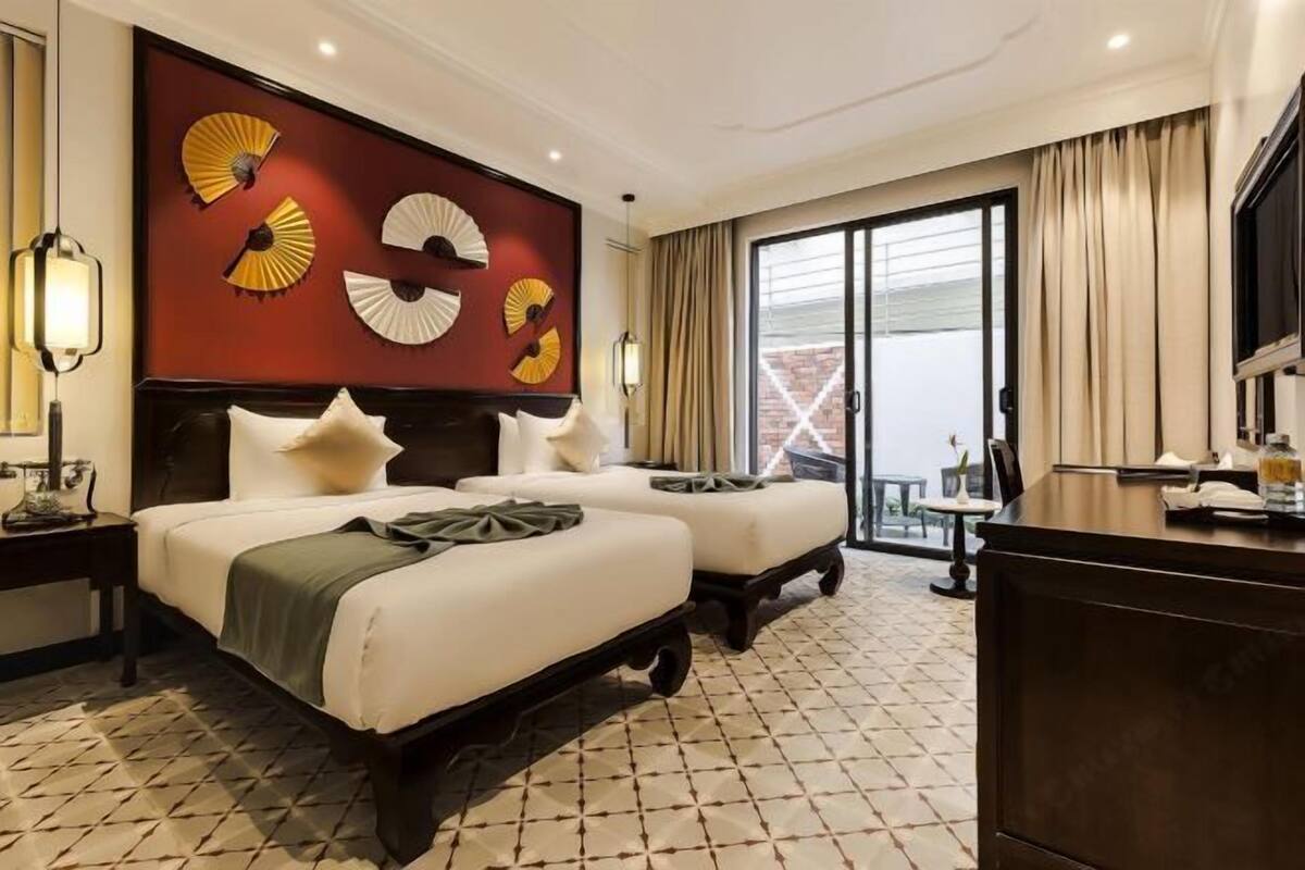 Habitación de hotel con cama, dos mesitas de noche, un escritorio, una silla, un ventanal grande con cortinas y una pared decorada con abanicos.