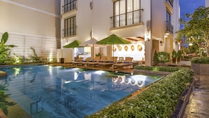 Zona de piscina con sillas de descanso y sombrillas, rodeada de follaje y un edificio con balcones.