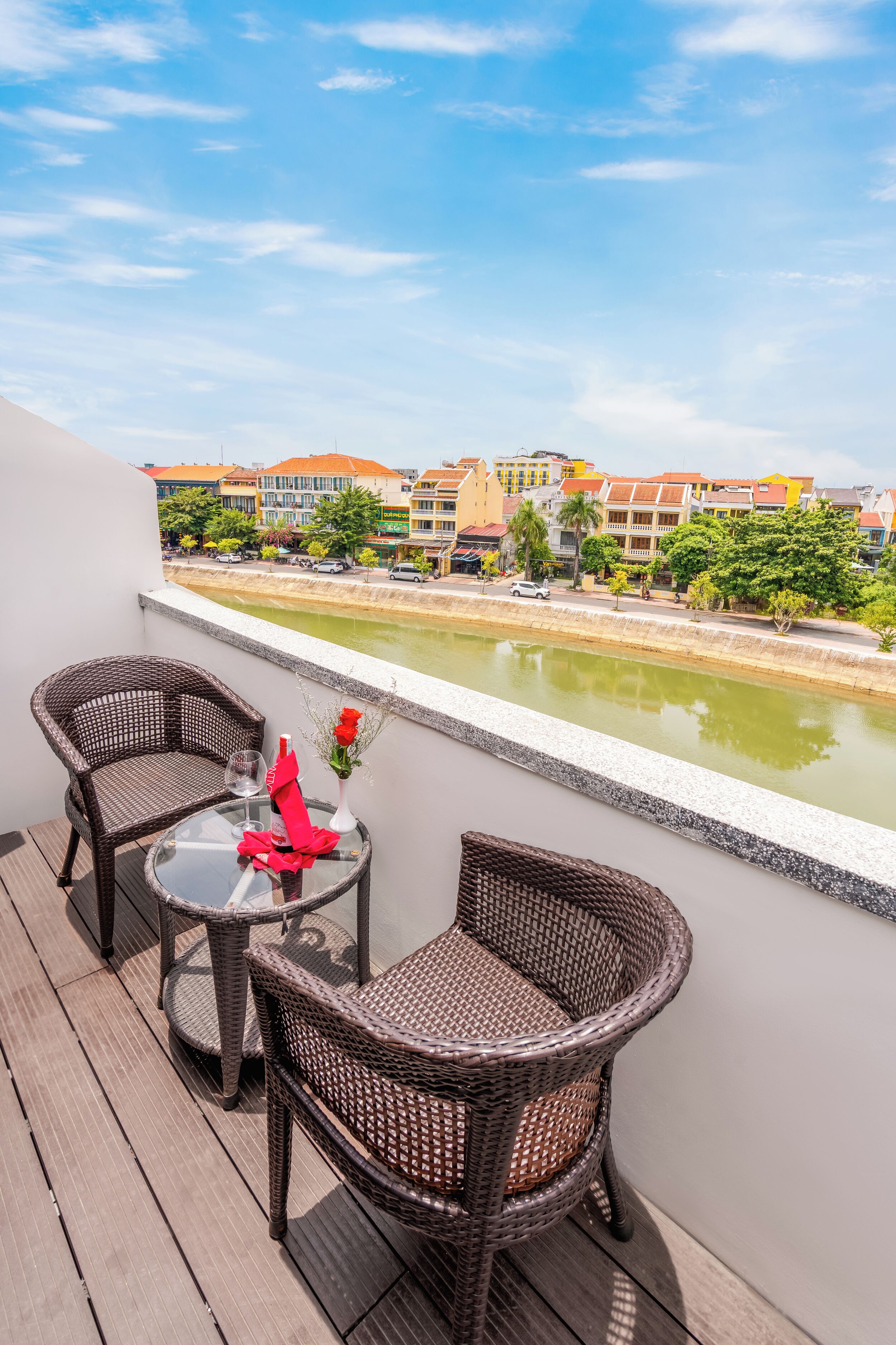 A balcony with wicker chairs and a table, overlooking a canal with buildings and greenery in the background.