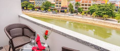 A balcony with wicker chairs and a table, overlooking a canal with buildings and greenery in the background.