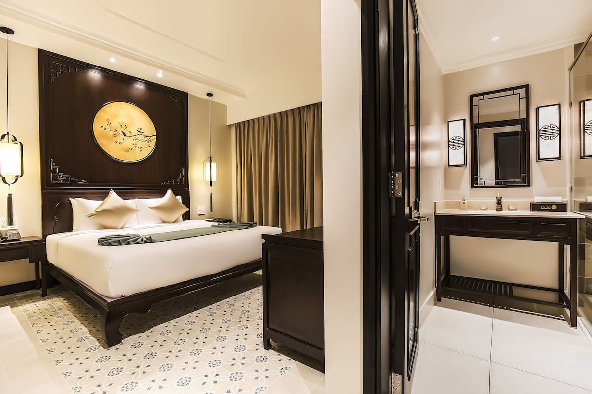 Una habitación de hotel con una cama grande, una mesita de noche de madera oscura, un espejo redondo fijado a la pared y un baño con tocador y espejo.
