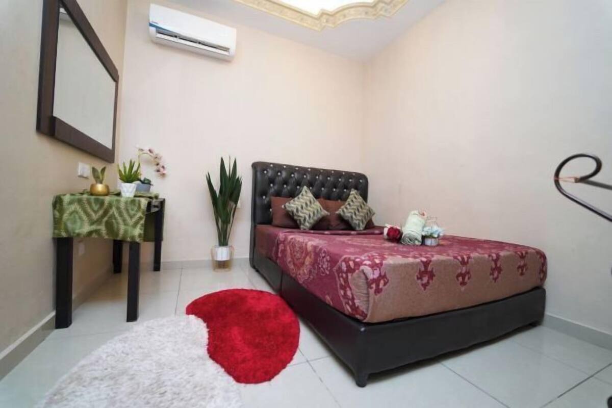 A bedroom with a bed, a nightstand, a potted plant, and a red rug.