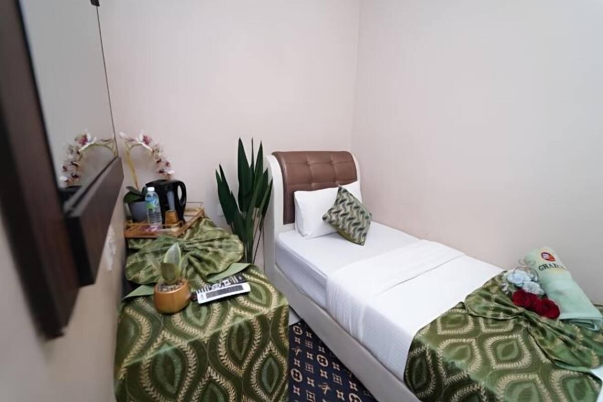 A small room with a bed, a bedside table, a potted plant, a mirror, and a wall-mounted shelf.