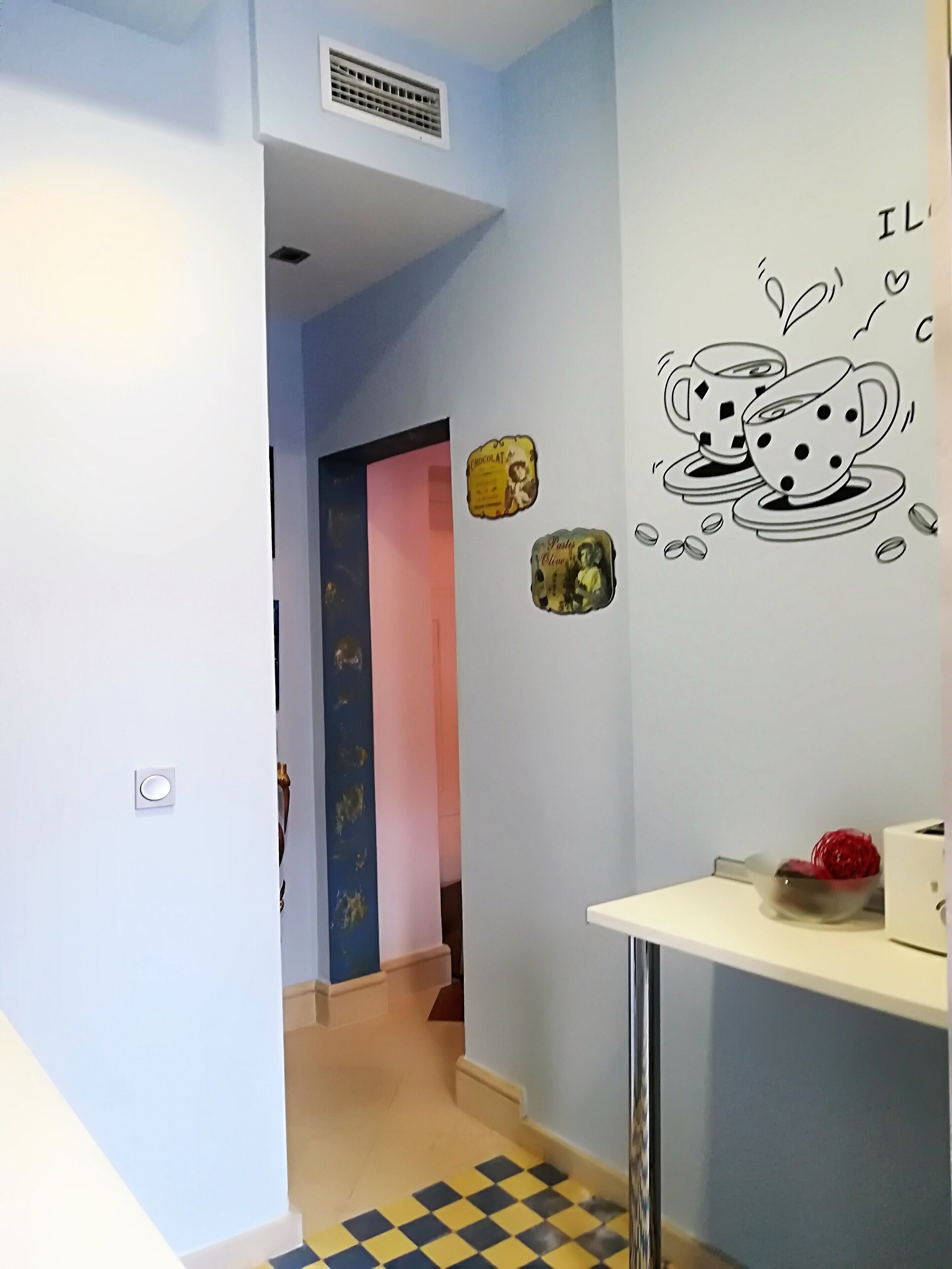 Un pasillo con piso a cuadros, una pared decorada con una taza de café y una puerta que da a otra habitación.