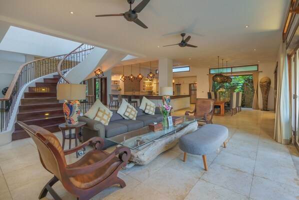 A spacious living area with a staircase, modern furniture, and a large window offering a view of the outdoors. - Villa Kaira (Canggu)