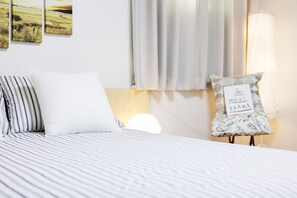A neatly made bed with striped bedding, a white pillow, and a pillow with a printed message. There are three framed pictures on the wall, a window with curtains, and a bedside lamp. - Tribear House 1 (Seoul)