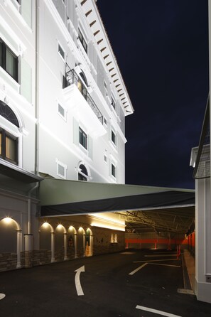 Entrada de un hotel con un corredor cubierto, zona de estacionamiento y fachada con varias ventanas y balcones.