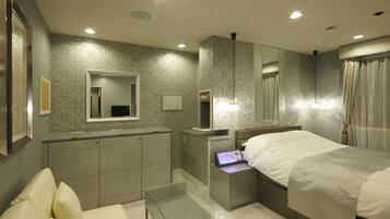 Una habitación de hotel moderna con una cama, una mesa de centro de vidrio, una zona de estar con sofá, un tocador con espejo y un baño con ducha.
