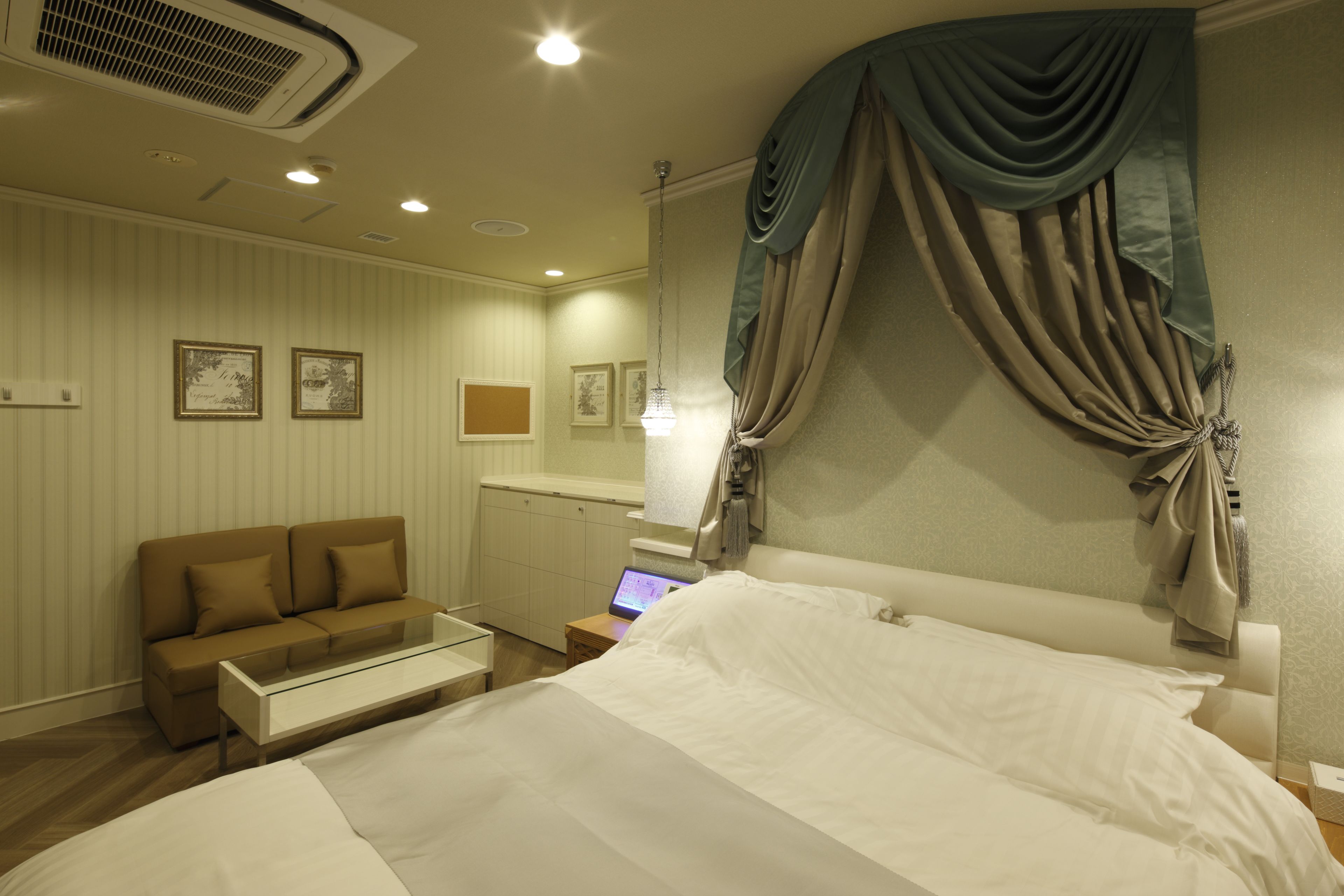 A hotel room with a bed, a sofa, a desk with a laptop, and a large window with curtains.