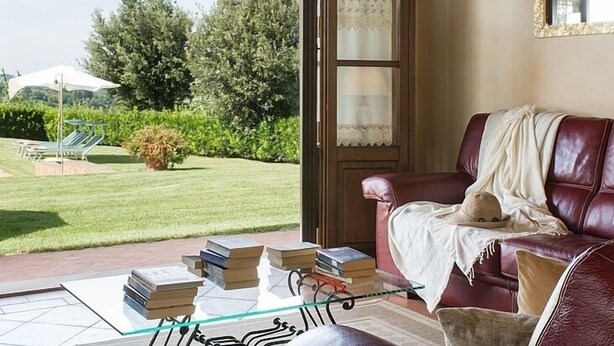 A room with a leather sofa, a glass coffee table, and a view of a garden through an archway. - Super panoramic, private pool, fast Wifi, Air cond, close to town - NO BILLS (Pisa)