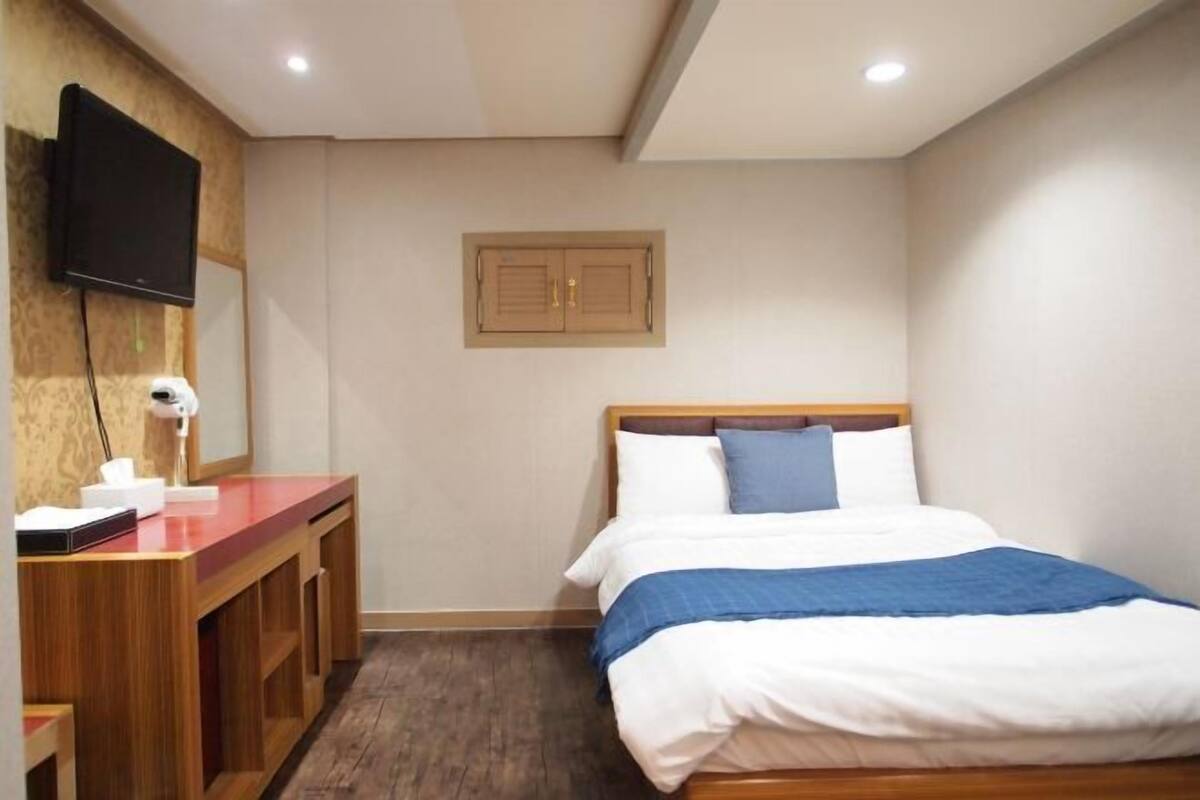 A hotel room with a bed, a desk with a flat-screen TV, a mirror, and a wooden door.