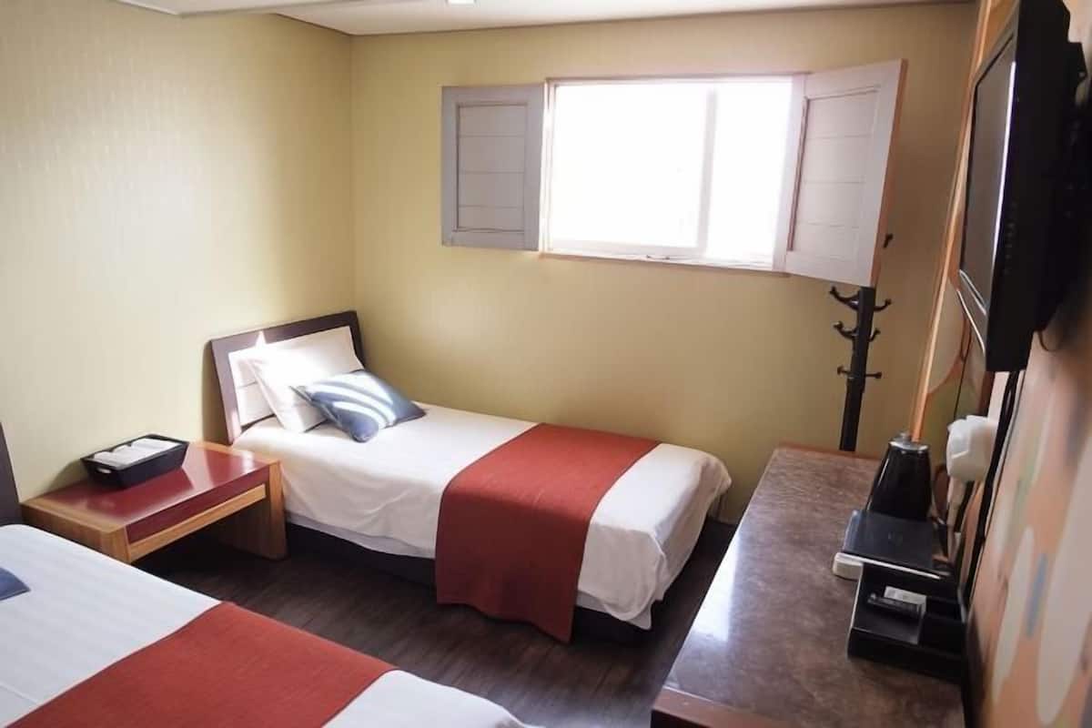 A small hotel room with two single beds, a desk, a chair, a TV, and a window with shutters.