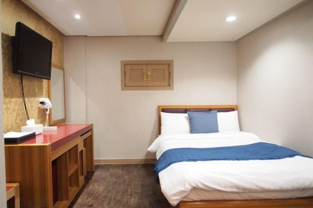 A hotel room with a bed, a desk with a flat-screen TV, a mirror, and a wooden door.