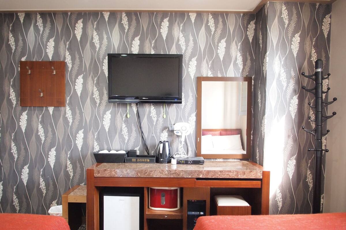 A hotel room with a bed, a TV mounted on the wall, a desk with a kettle, and a mirror.