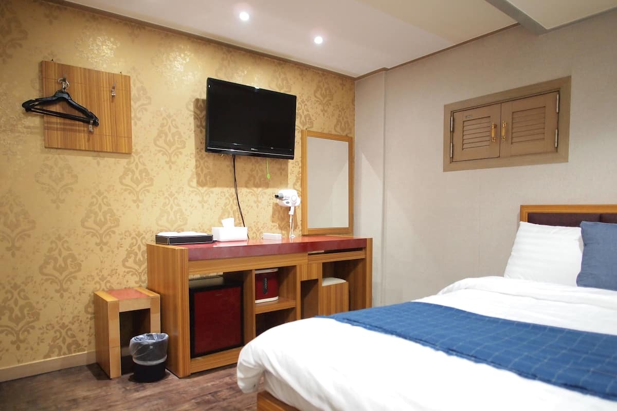 A hotel room with a wooden desk, a flat-screen TV, a mirror, a bed with white and blue bedding, and a trash bin.