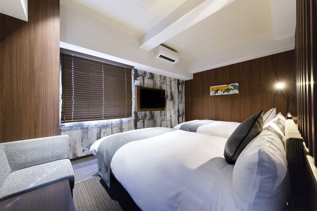A hotel room with two beds, a sofa, a flat-screen TV mounted on the wall, and a window with blinds.