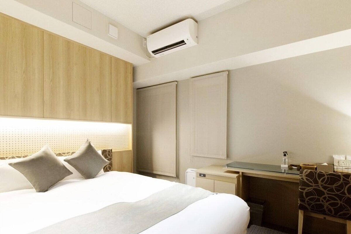 A hotel room with a bed, a desk, a chair, an air conditioning unit, and a window with blinds.