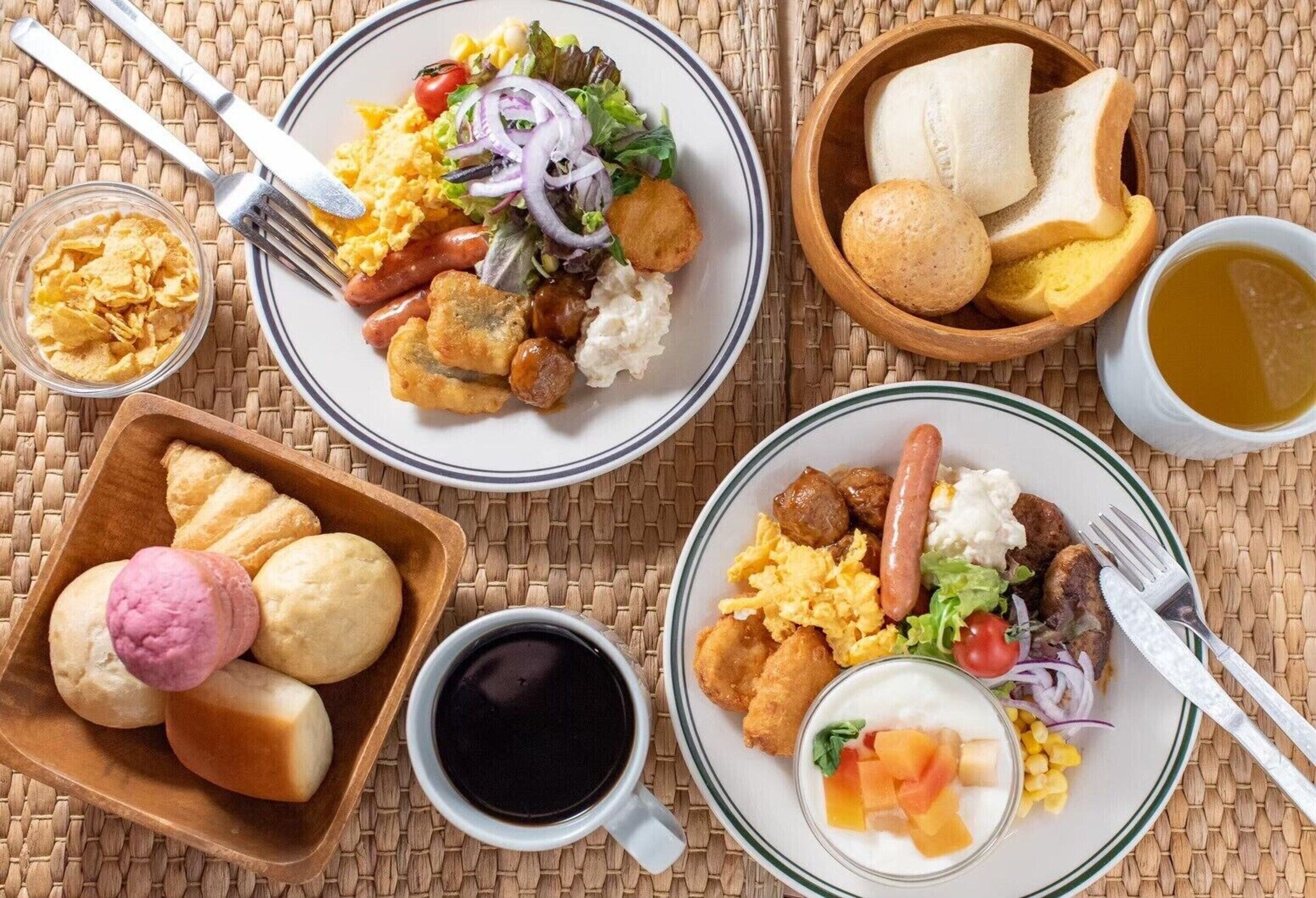 A breakfast setting with plates of food, including scrambled eggs, sausages, and a salad, along with bread, a bowl of chips, and a cup of coffee.