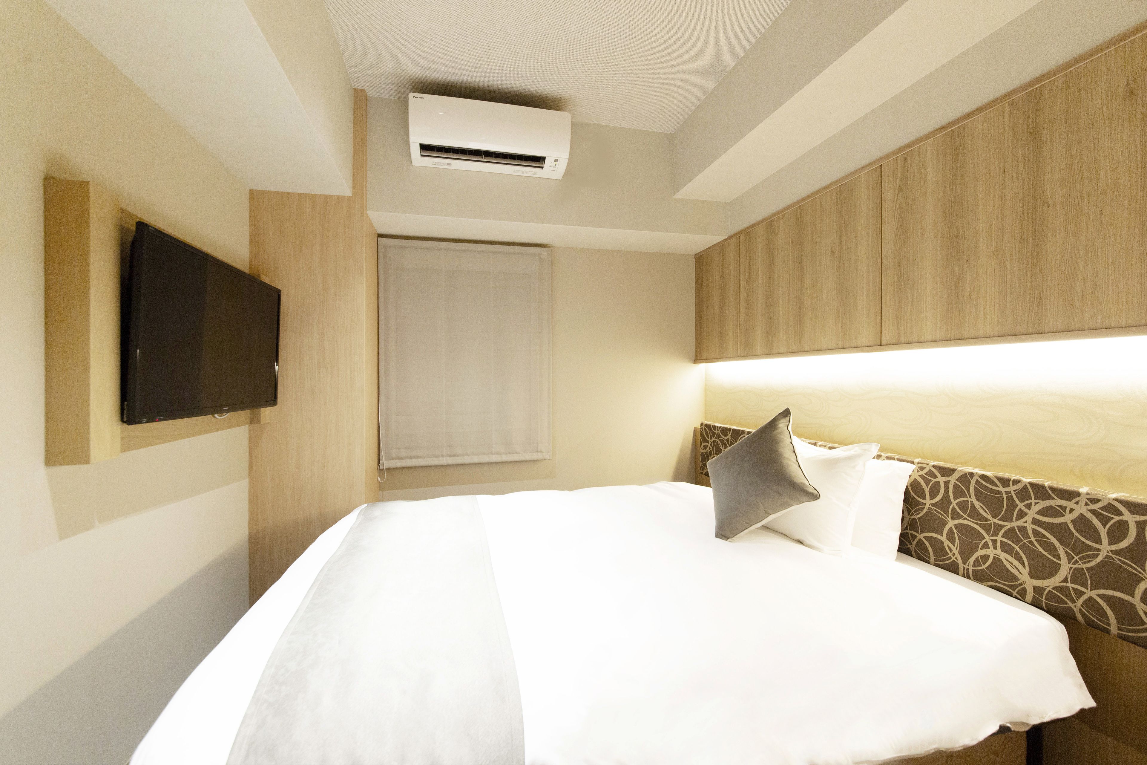 A hotel room with a bed, a flat-screen TV mounted on the wall, and an air conditioning unit above the bed.
