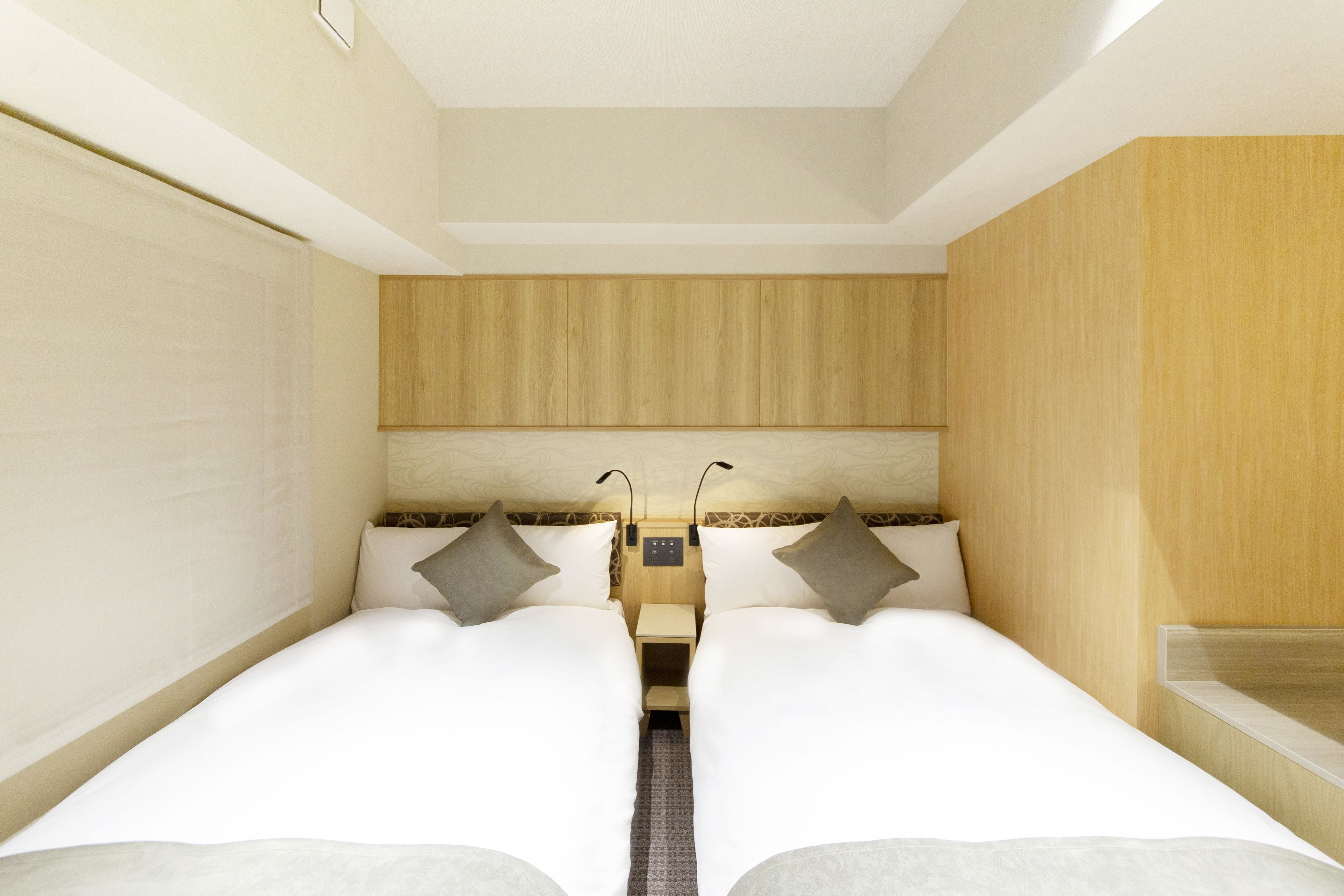 Habitación de hotel pequeña con dos camas individuales, una mesita de noche con lámpara y cabecera de madera.
