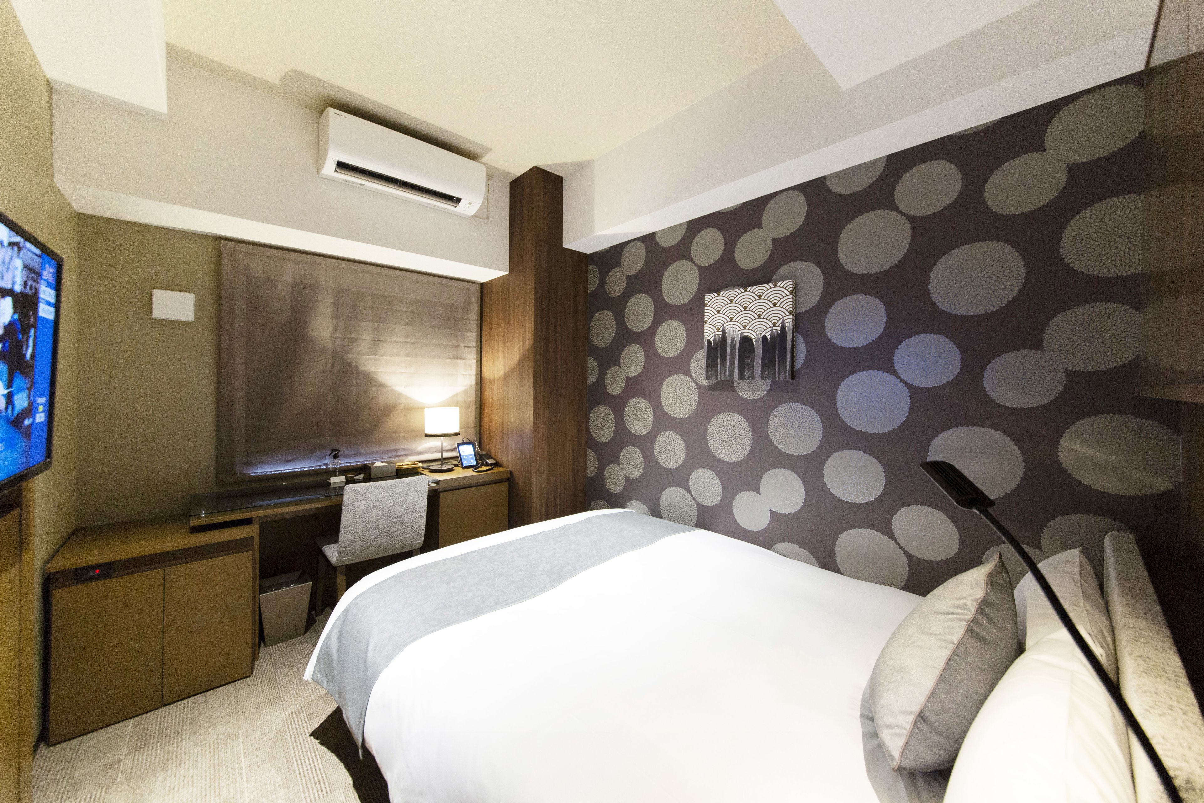 A hotel room with a bed, a desk, a chair, an air conditioner, and a television.