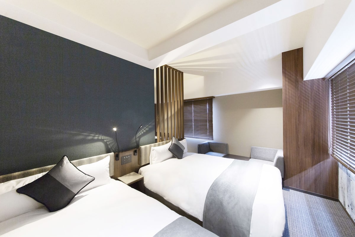 A modern hotel room with two beds, a large window with blinds, a dark accent wall, and a wooden panel on the right.