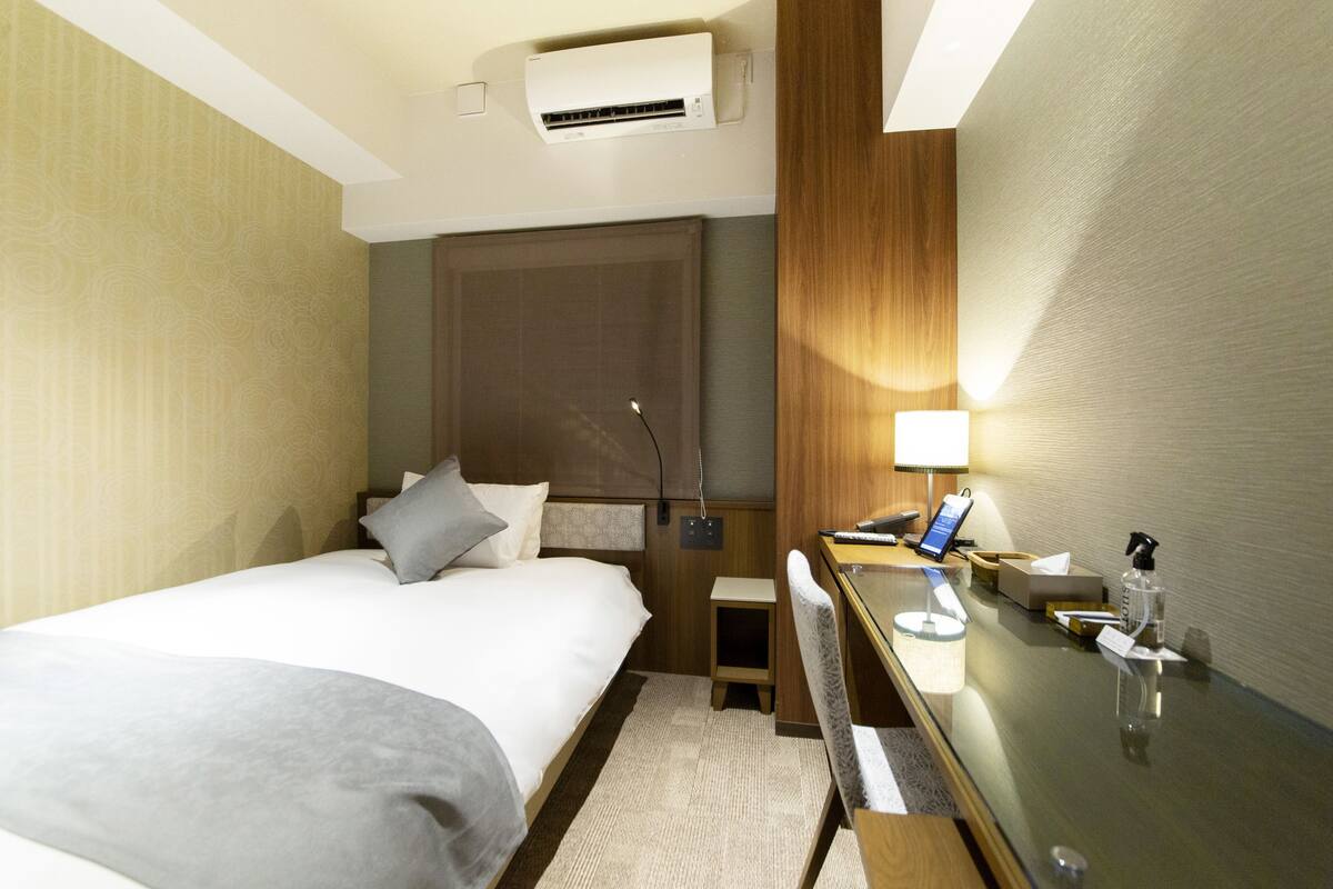 A hotel room with a bed, bedside table, desk, chair, and a desk lamp.