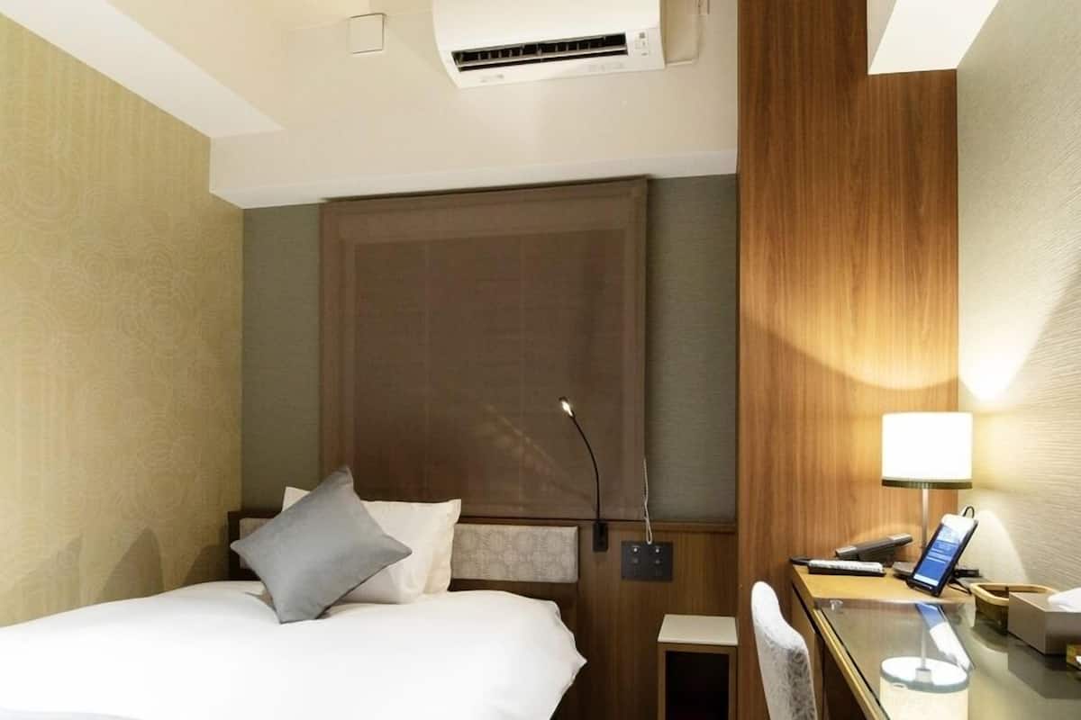 A hotel room with a bed, bedside table, lamp, and an air conditioning unit.