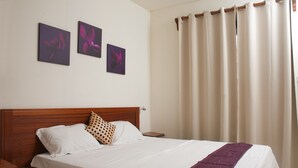 A neatly made bed with a wooden headboard, a white pillow, a patterned pillow, and a purple throw. There are three framed artworks on the wall above the bed.
