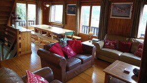 A cozy cabin interior with a wooden dining table, leather couches, and large windows offering views of the outdoors.