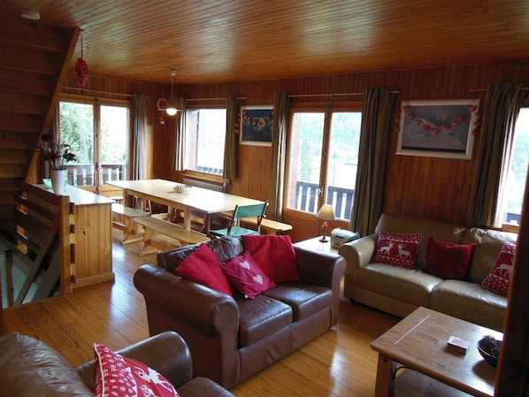 L’intérieur chaleureux d’un chalet avec une table à manger en bois, des canapés en cuir et de grandes fenêtres offrant une vue sur le paysage extérieur.