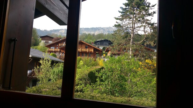 A view of a wooden cabin with a balcony, surrounded by lush greenery and a mountainous backdrop.